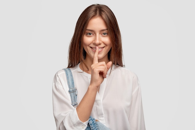 Shh, its privacy! Good looking dark haired woman with charming smile, keeps index finger over mouth