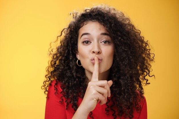 Shh please not tell secret. Cute and tender woman with curly hairstyle gently shushing with index finger on folded lips asking keep promise and remain silent, making surprise over yellow wall.