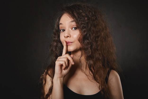 Shh. Don't tell anyone. Picture of emotional funny brunette girl in tank top keeping fore ginger at her lips, raising eyebrows, saying Shh, asking not to disclose her secret. Confidential information