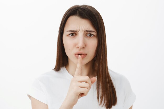 Shh you promise stay quiet. Portrait of displeased and upset cute brunette in 20s frowning disappointed holding index finger over mouth as asking keep secret with shush gesture over gray wall