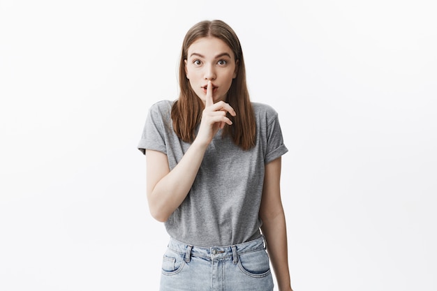 Shhh. close up di bella giovane ragazza bruna allegra con capelli klong e occhi castani in abiti grigi casual tenendo il dito davanti alla bocca, con espressione gentile, giocando con