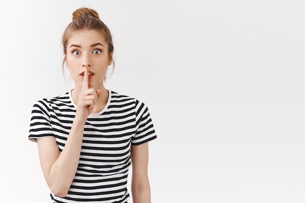 Shhh dont say word. Excited young silly girl with bun wear striped t-shirt, press finger to lips seal with promise not tell anybody, hiding secret, gossiping behind coworker back, white background