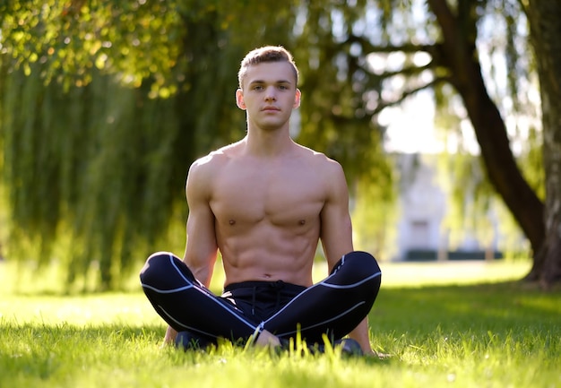 Молодой мужской фитнес-модель без рубашки сидит на траве в парке.