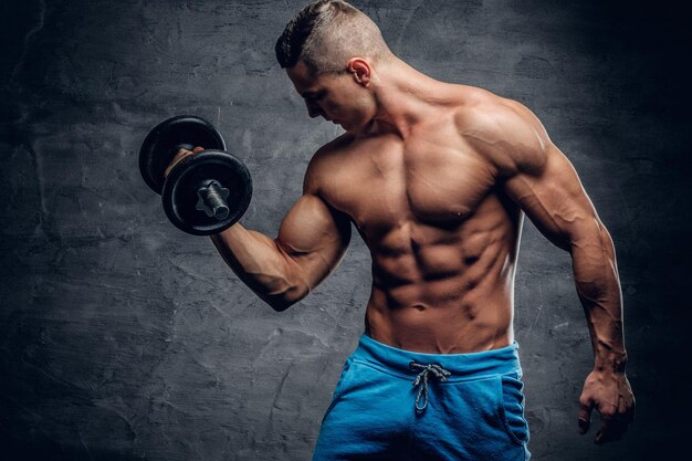 Shirtless athletic male doing biceps workouts with one dumbbell on grey vignette background.