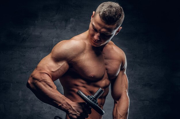 Shirtless athletic male doing biceps workouts with one dumbbell on grey vignette background.