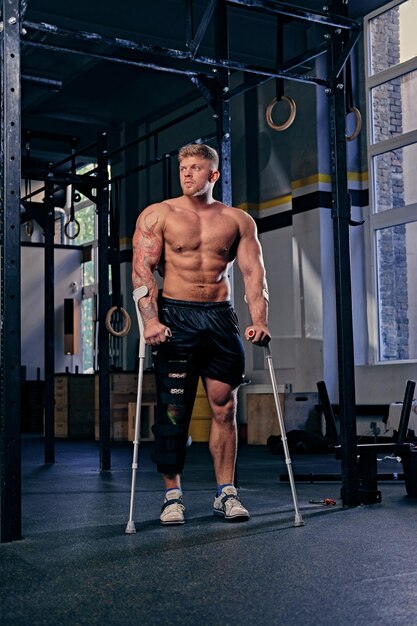 Shirtless bodybuilder on crutches near cross fit stand in a gym.