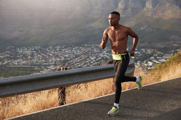 Shirtless dark skinned man runs quickly along road, wears sportshoes, has motivation to be winner of race competitions