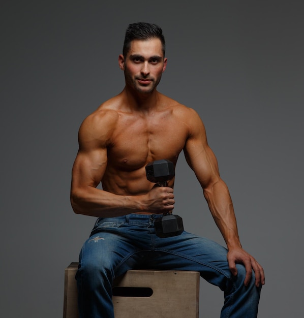 Shirtless fitness man in blue jeans sitting on podium and holding dumbell. Isolated on grey background