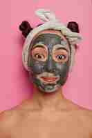 Free photo shirtless glad asian woman applies black beauty mask, enjoys anti wrikle or anti puffiness procedures, spa treatments, has two combed buns, wears headband, isolated over rosy  wall