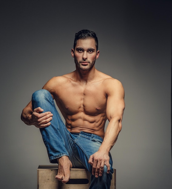 Shirtless guy in blue jeans sitting on podium. Isolated on grey background.