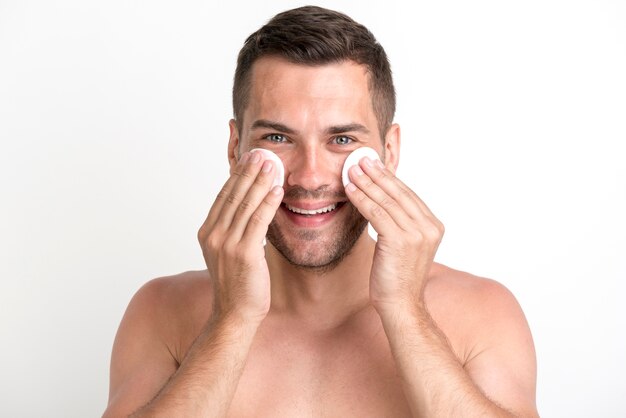 Shirtless man cleaning his face with batting cotton pads over white background and looking at camera