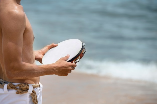 Shirtless man practicing capoeira by on the beach with tambourine