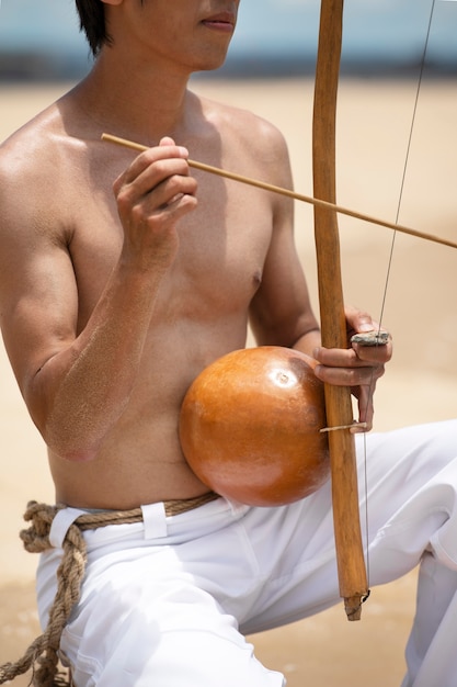 Free photo shirtless man practicing capoeira by on the beach with wooden bow