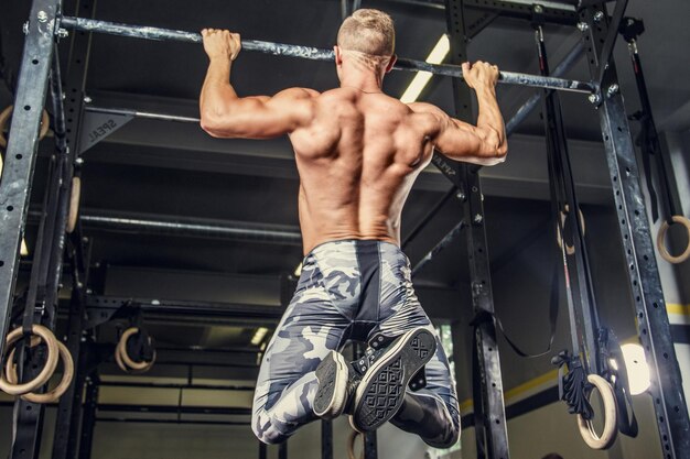 Shirtless man pulling up on horizontal bar in a gym.