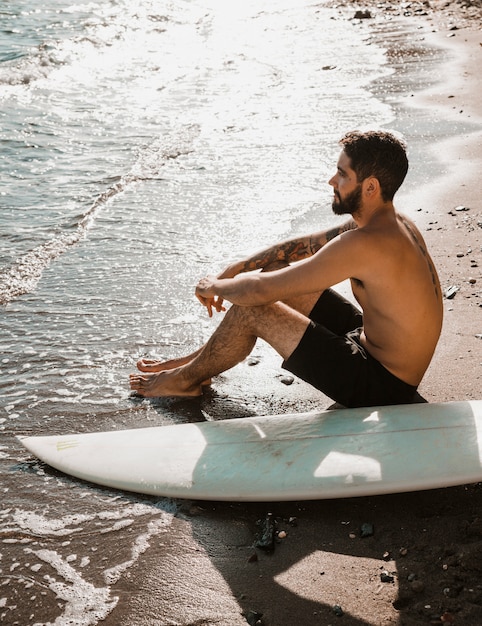 Foto gratuita uomo senza camicia con tavola da surf rilassante vicino al mare agitando