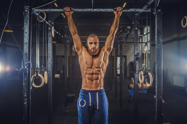 Shirtless muscular bearded male doing exercises on horizontal bar in a gym club.