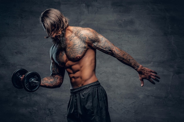 Foto gratuita maschio muscoloso e barbuto senza camicia con un tatuaggio sul busto che fa un allenamento bicipite con manubrio su sfondo grigio scuro.