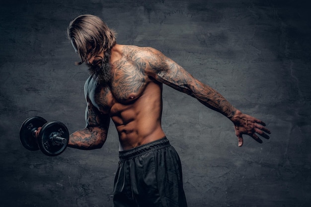 Shirtless muscular, bearded male with a tattoo on his torso doing a biceps workout with dumbbell over dark grey background.