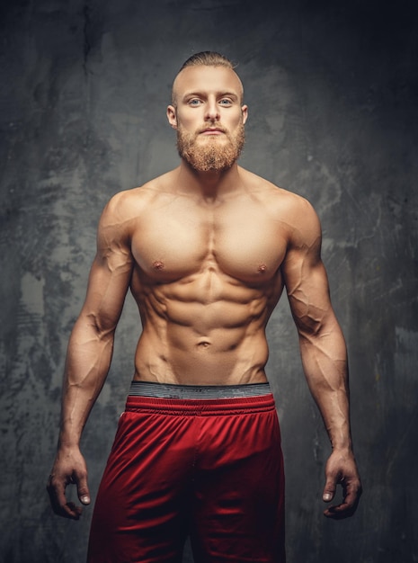 Shirtless muscular bearded man in red pants posing in studio over grey background.