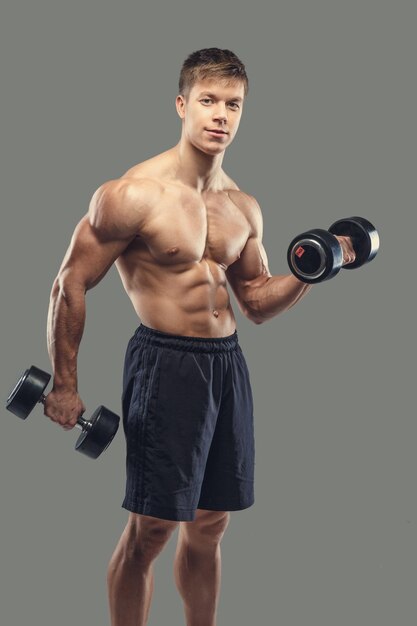 Shirtless muscular male doing biceps exercises with dumbbells on a grey background.