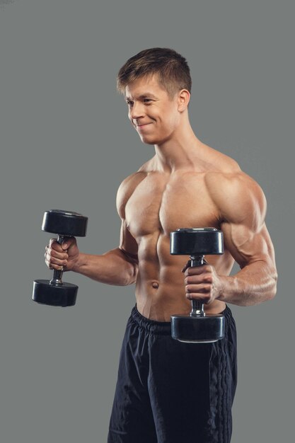 Shirtless muscular male doing biceps exercises with dumbbells on a grey background.