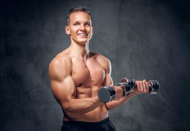 The shirtless muscular male holds a set of dumbbells over dark grey vignette background.