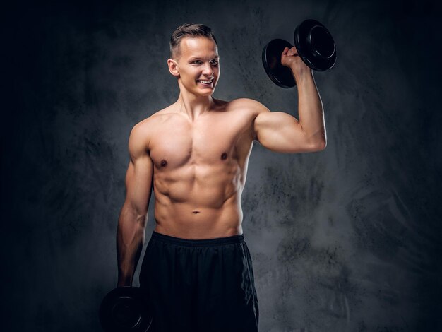 The shirtless muscular male holds a set of dumbbells over dark grey vignette background.