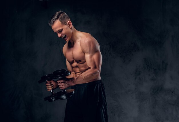 The shirtless muscular male holds a set of dumbbells over dark grey vignette background.