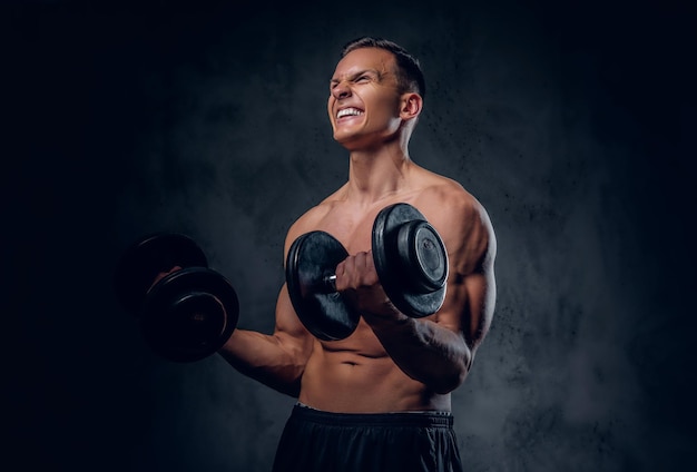 The shirtless muscular male holds a set of dumbbells over dark grey vignette background.