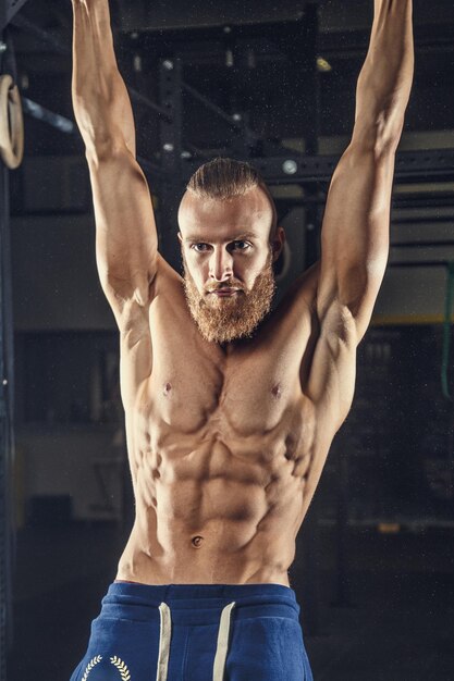 Shirtless muscular man in blue sports pants doing exercises in a gym on horizontal bar.