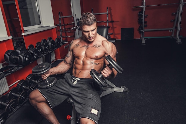 Shirtless muscular man doing biceps workout with dumbbells in a gym club.