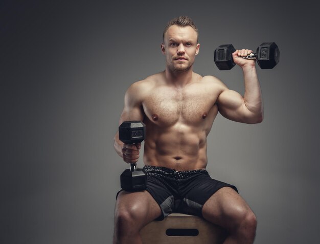 Shirtless muscular man holds a pair of dumbbells.