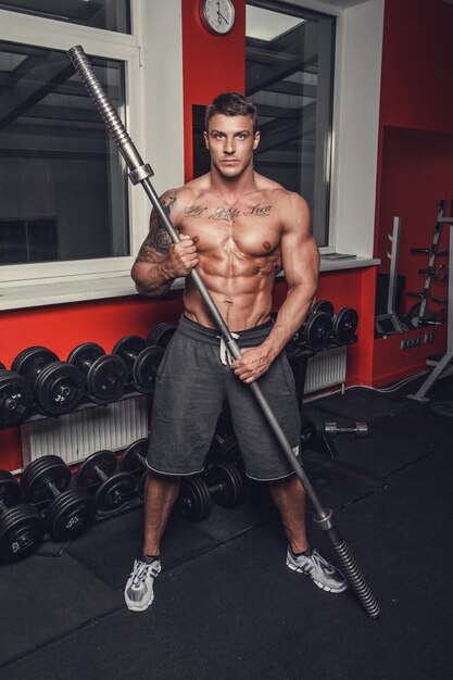 Shirtless muscular tattooed man holds barbell in a gym club.