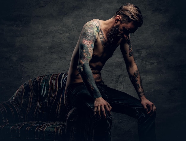 Free photo the shirtless, tattooed bearded male sits on a chair over grey background.