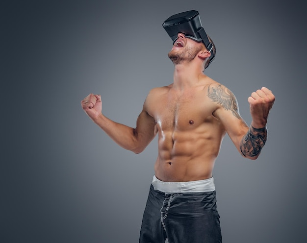 A shirtless, tattooed fighter with virtual reality glasses on his head isolated on grey background.