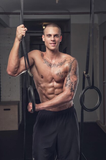 Free photo shirtless tattooed man doing ring workout in a gym club.