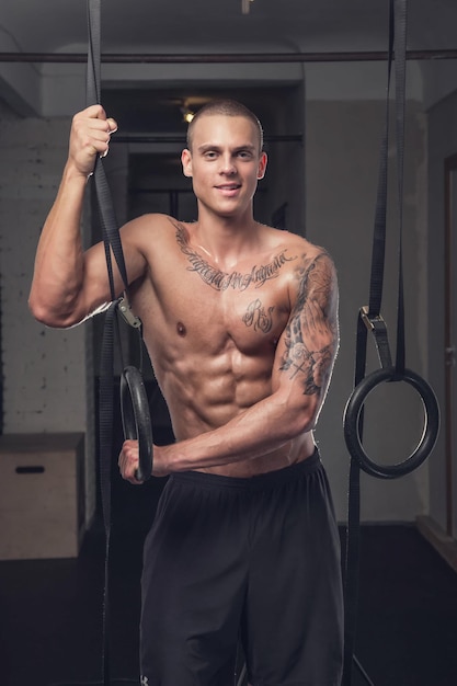 Shirtless tattooed man doing ring workout in a gym club.