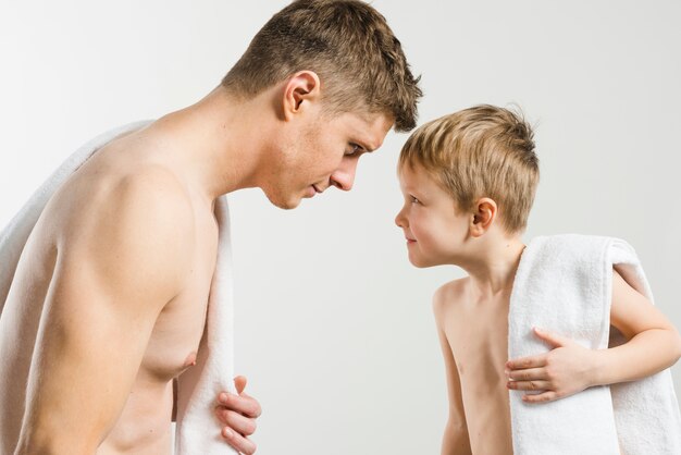 Shirtless young man and his son with folded towel on their shoulder looking at each other against grey backdrop