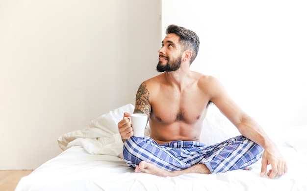 Free photo shirtless young man sitting on bed holding cup of coffee