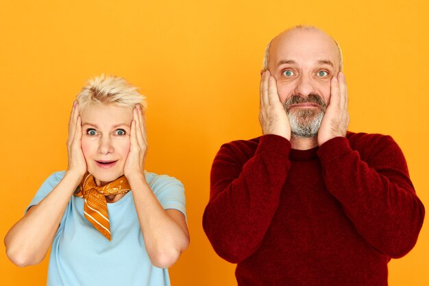 Shock, astonishment and surprise concept. Funny European bearded senior male and blonde female being shocked with amazing unexpected news, holding hands on cheeks, keeping mouth wide opened