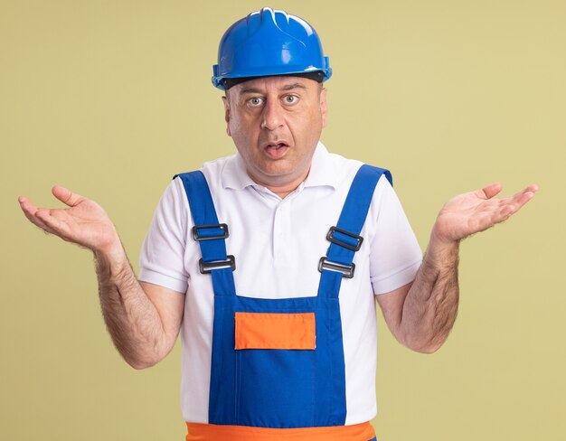 Shocked adult builder man in uniform holds hands open isolated on olive green wall