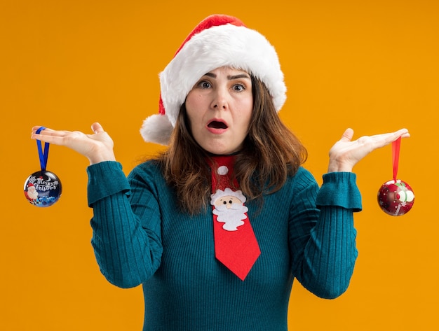 Shocked adult caucasian woman with santa hat and santa tie holding glass ball ornaments isolated on orange background with copy space