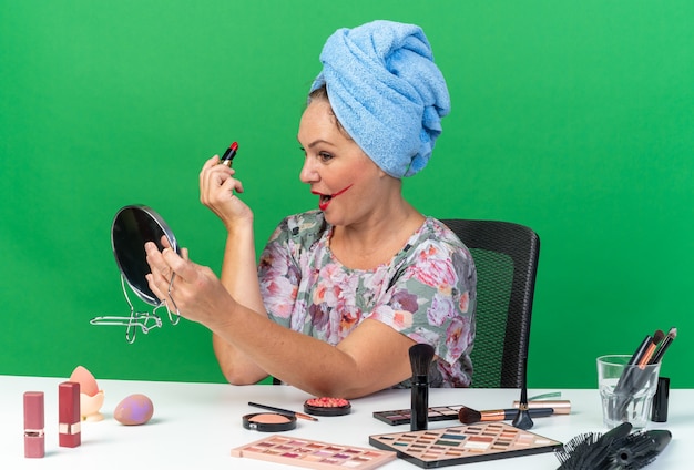 Shocked adult caucasian woman with wrapped hair in towel sitting at table with makeup tools holding lipstick and looking at mirror isolated on green wall with copy space