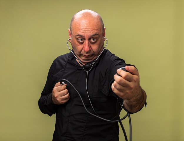 shocked adult ill caucasian man holding and looking at sphygmomanometer isolated on olive green wall with copy space