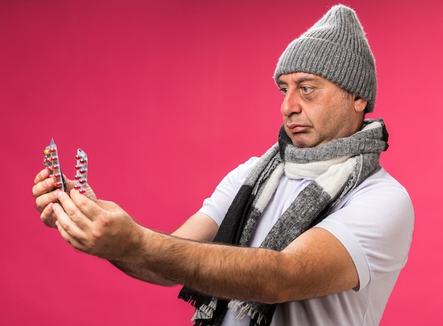 Uomo caucasico malato adulto scioccato con sciarpa intorno al collo che indossa cappello invernale tenendo e guardando diversi pacchetti di medicinali isolati sul muro rosa con spazio di copia