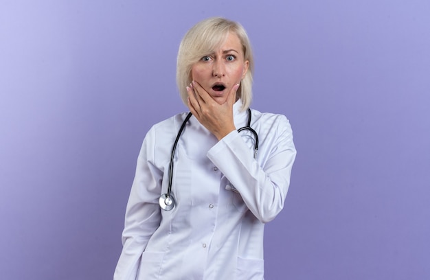 청진 기 들고 턱 복사 공간이 보라색 벽에 고립 된 의료 가운에 충격 된 성인 슬라브 여성 의사
