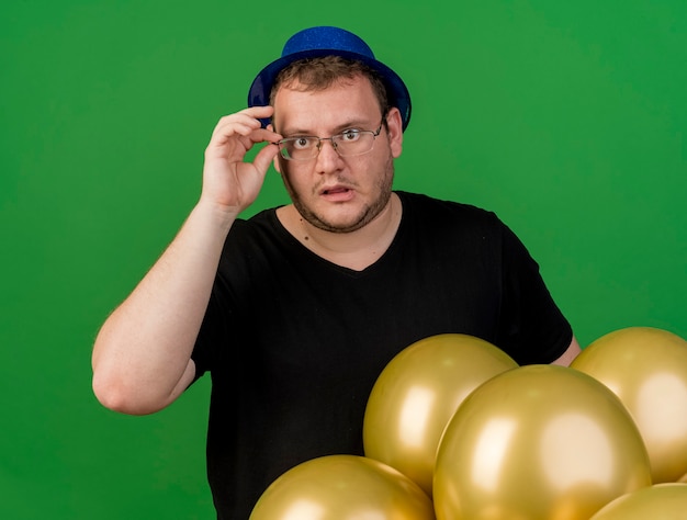 Shocked adult slavic man holds and looks through optical glasses at camera wearing blue party hat standing with helium balloons 