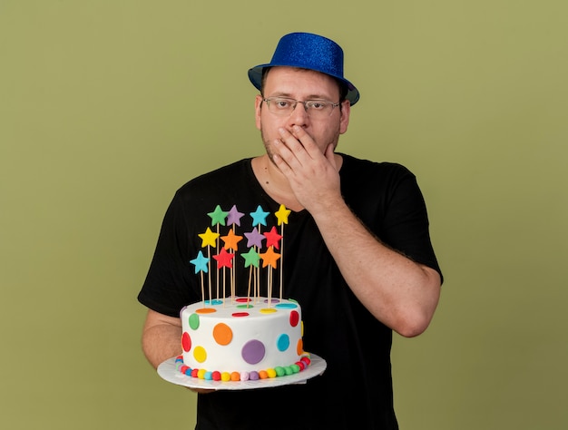 Foto gratuita scioccato adulto slavo con occhiali ottici che indossa un cappello da festa blu mette la mano sulla bocca e tiene la torta di compleanno