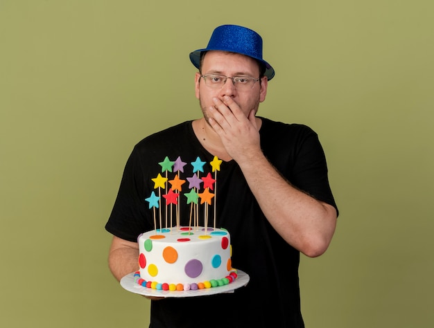 파란색 파티 모자를 쓰고 광학 안경에 충격을받은 성인 슬라브 남자가 입에 손을 대고 생일 케이크를 들고