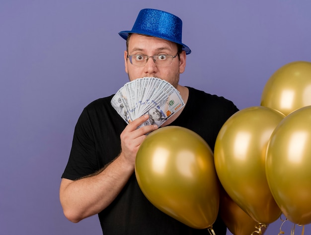 Foto gratuita uomo slavo adulto scioccato con occhiali ottici che indossa un cappello da festa blu con palloncini di elio con denaro